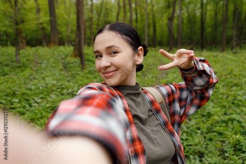 Smiling female hiker wearing red plaid shirt takes selfie and makes peace sign while exploring lush green forest, enjoying peaceful nature and sharing her adventure