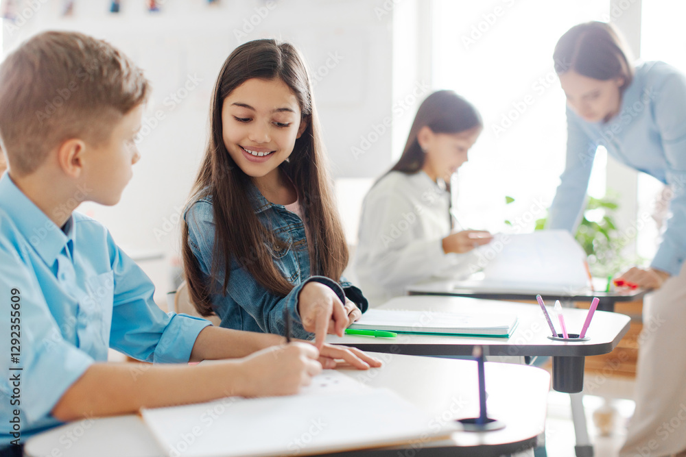 European schoolgirl helping her classmate boy and pointing at his copybook, sitting at desks in classroom during lesson in private school