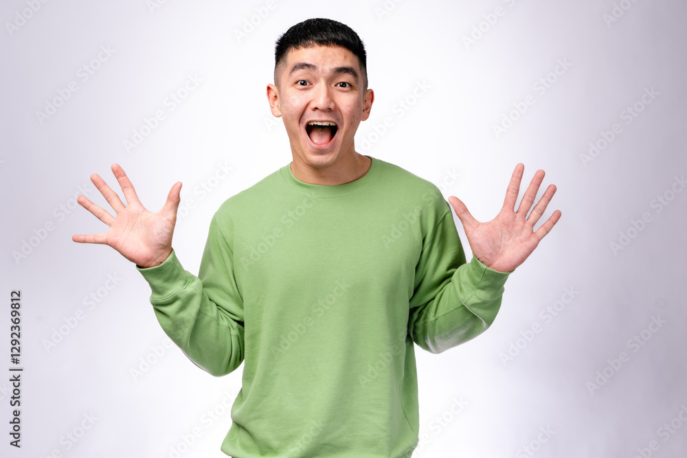 An excited Asian man in a green sweatshirt raises both hands with wide-open palms and a joyful expression. He stands against a plain white background, conveying surprise, excitement An excited Asian man in a green sweatshirt raises both hands with wide-open palms and a joyful expression. He stands against a plain white background, conveying surprise, excitement