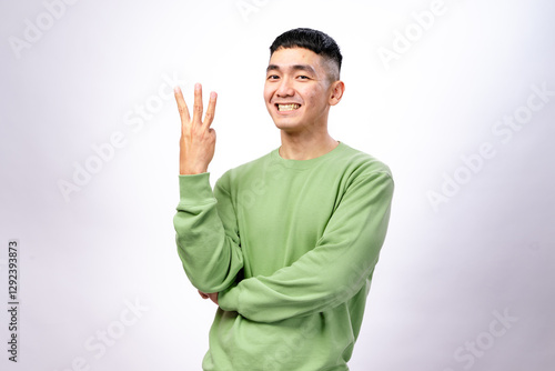 An Asian man in a green sweatshirt is smiling while holding up three fingers on his right hand. His left arm is crossed, and he stands against a white background, expressing confidence and enthusiasm
