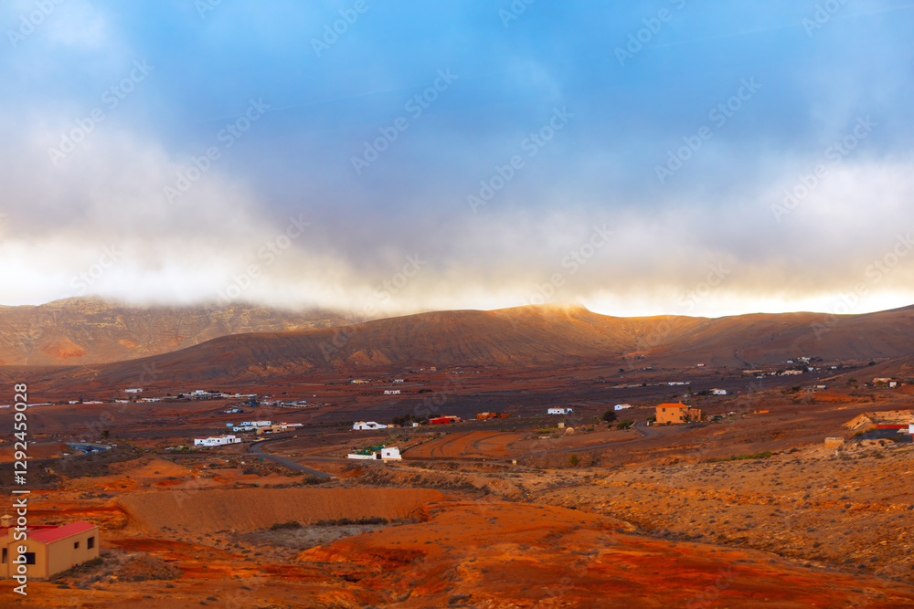 Rugged and arid landscape of Fuerteventura, with scattered houses and buildings dotting the terrain. Canary Islands scene is dominated by rolling hills and a dramatic sky filled with thick clouds