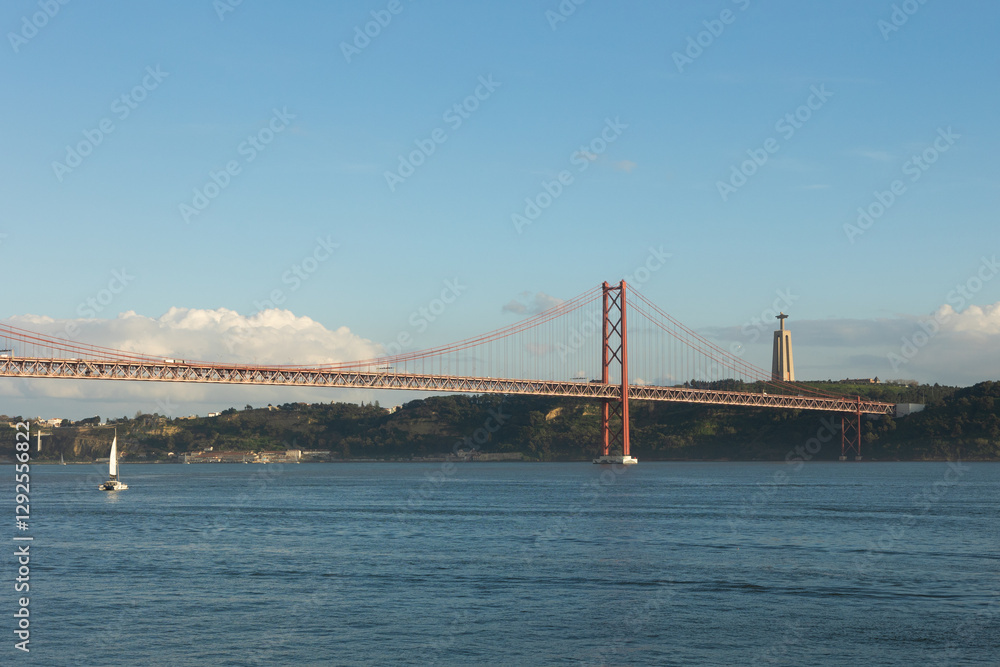 Naklejka premium A stunning view of the 25 de Abril Bridge in Lisbon, Portugal, spanning the Tagus River with the Cristo Rei monument in the background. A sailboat glides through the water under a clear blue sky.