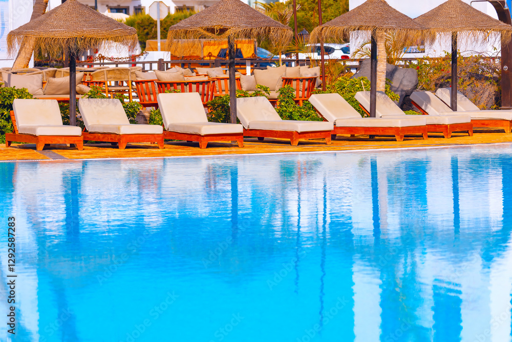 Poolside area with beige lounge chairs arranged in a row under thatched umbrellas, reflecting in blue water. Setting appears to be relaxing outdoor space of resort, designed for leisure and comfort