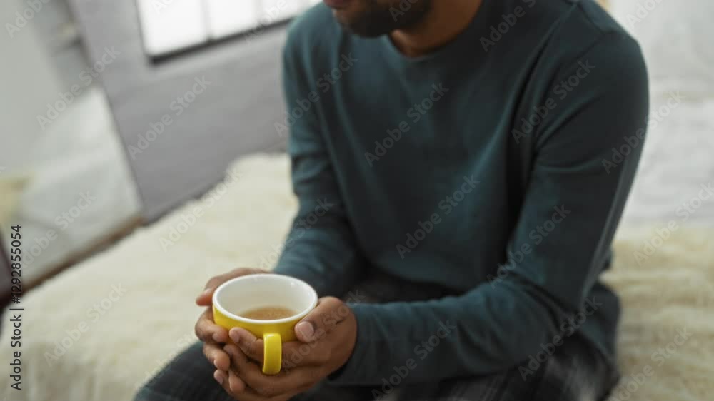 Young man with beard sitting on bed holding yellow cup in modern bedroom with window, wearing casual pajamas and green sweater, reflecting a relaxed, cozy morning vibe. Young man with beard sitting on bed holding yellow cup in modern bedroom with window, wearing casual pajamas and green sweater, reflecting a relaxed, cozy morning vibe.