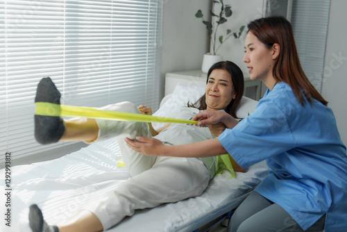Asian physiotherapist helping patient doing exercise with resistance band in hospital bed