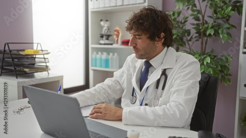 Wallpaper Mural Young male doctor working on a laptop in a bright hospital office with medical equipment, files, and a stethoscope, capturing a thoughtful and focused professional environment. Torontodigital.ca