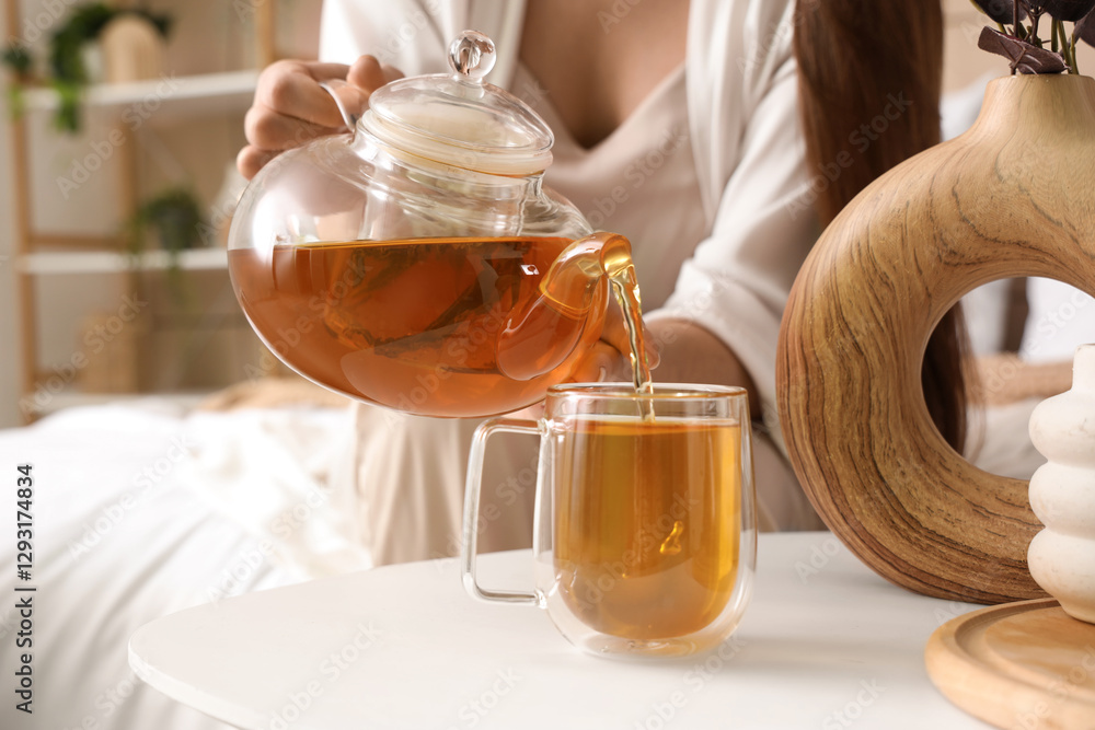 Young woman pouring green tea into glass cup on table in bedroom, closeup Young woman pouring green tea into glass cup on table in bedroom, closeup