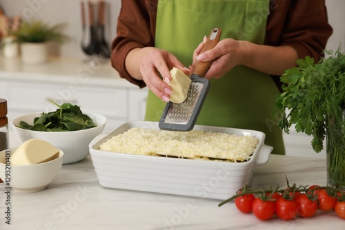 Woman grating cheese onto spinach lasagna at marble table indoors, closeup