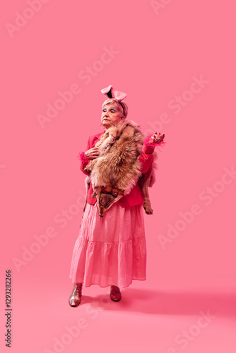 Fotografie Elderly woman in pink dress, furry stole, and bunny ears standing against monochrome pink background