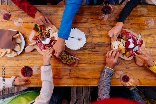 Friends sharing a rustic meal together outdoors - group of people enjoying a shared meal with wine, cheese, bread, and charcuterie on a wooden table