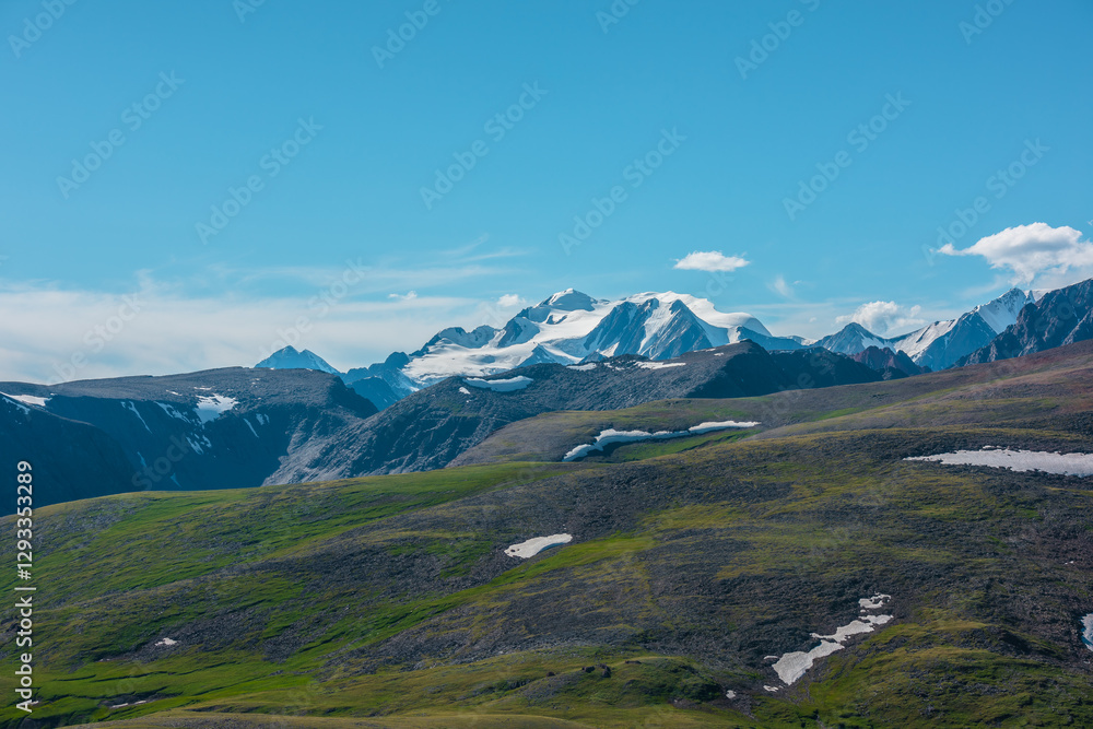 Fototapeta premium Aerial top view to big snow castle in bright sun under clouds in blue sky. Scenic landscape with sunlit green hills with snowfields against large snow-white rocky mountain peak far away in sunny day.