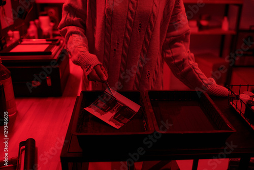 Close up of young woman dipping developed photo print in chemical tray while working on analog prints in darkroom lit by red light copy space