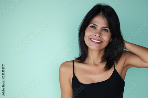 Studio portrait of a beautiful smiling mid-adult woman with dark hair wearing a black top, touching her hair and posing against a turquoise background
