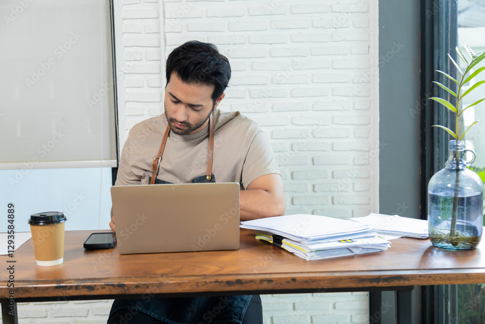 Fototapeta premium Attractive barista male or employee concentrate on stacks of tax declaration bills, restaurant owner using laptop for bookkeeping control payment, multitask small business lifestyle doing everything