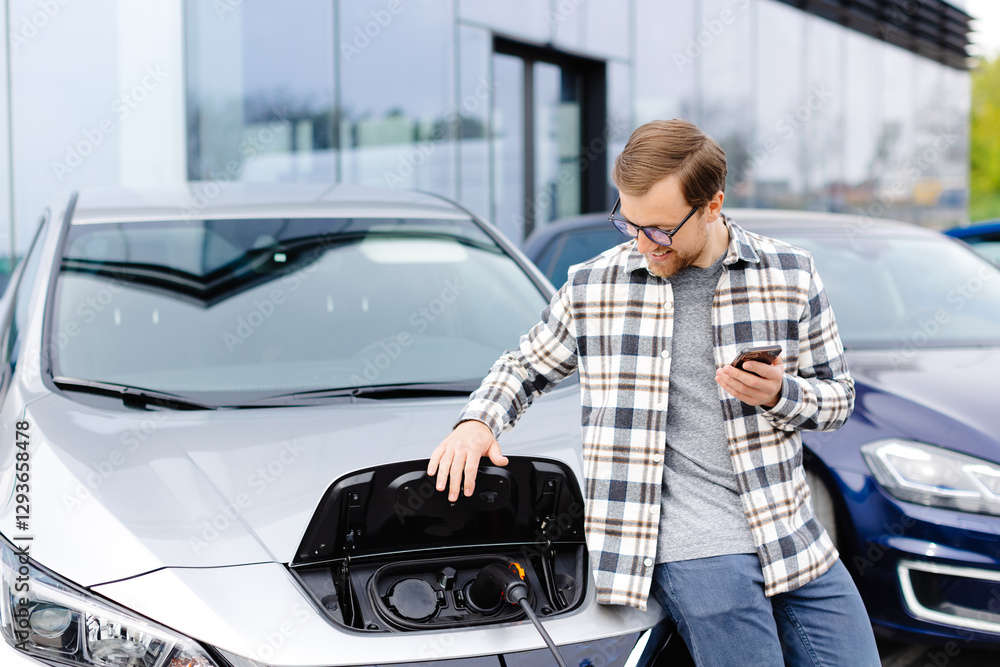 A young man uses a mobile phone while leaning on his electric car while the car is charging A young man uses a mobile phone while leaning on his electric car while the car is charging
