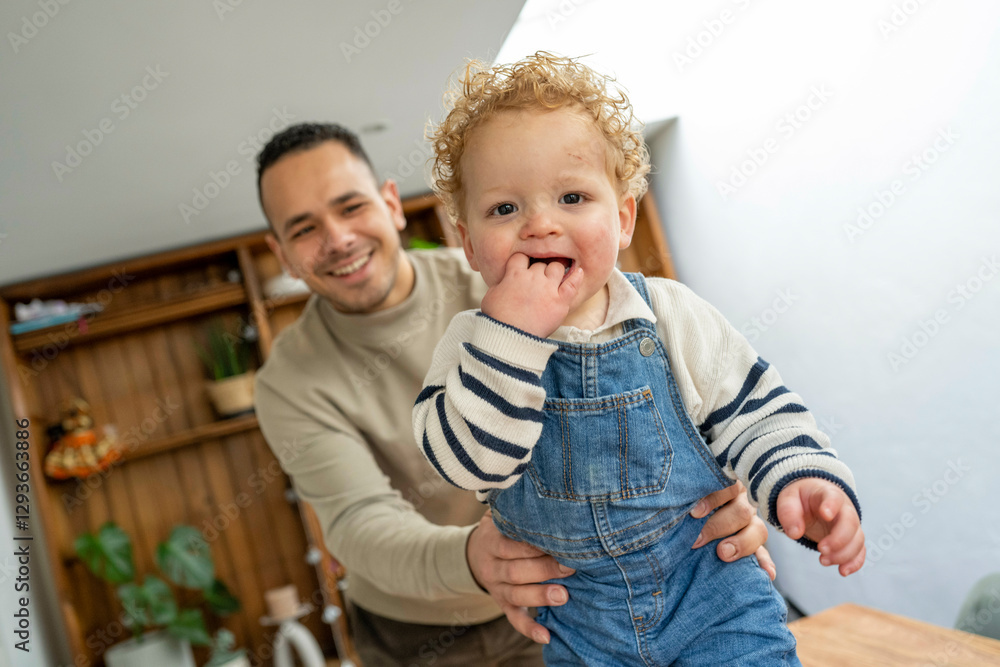 Joyful toddler with curly hair wearing denim overalls, chewing on his finger, with a smiling father in the background, Netherlands Joyful toddler with curly hair wearing denim overalls, chewing on his finger, with a smiling father in the background, Netherlands