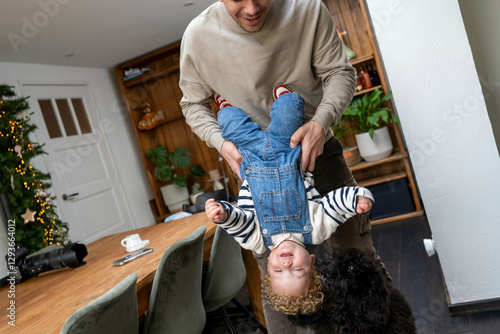 Father playing with a laughing child by holding them upside down as a black dog joins the fun in a cozy home kitchen, Netherlands photo