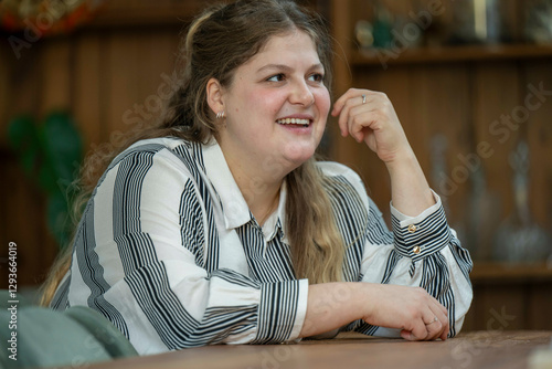 Smiling young woman resting her chin on her hand at a wooden table, looking away with a joyful expression in a casual indoor setting, Netherlands photo