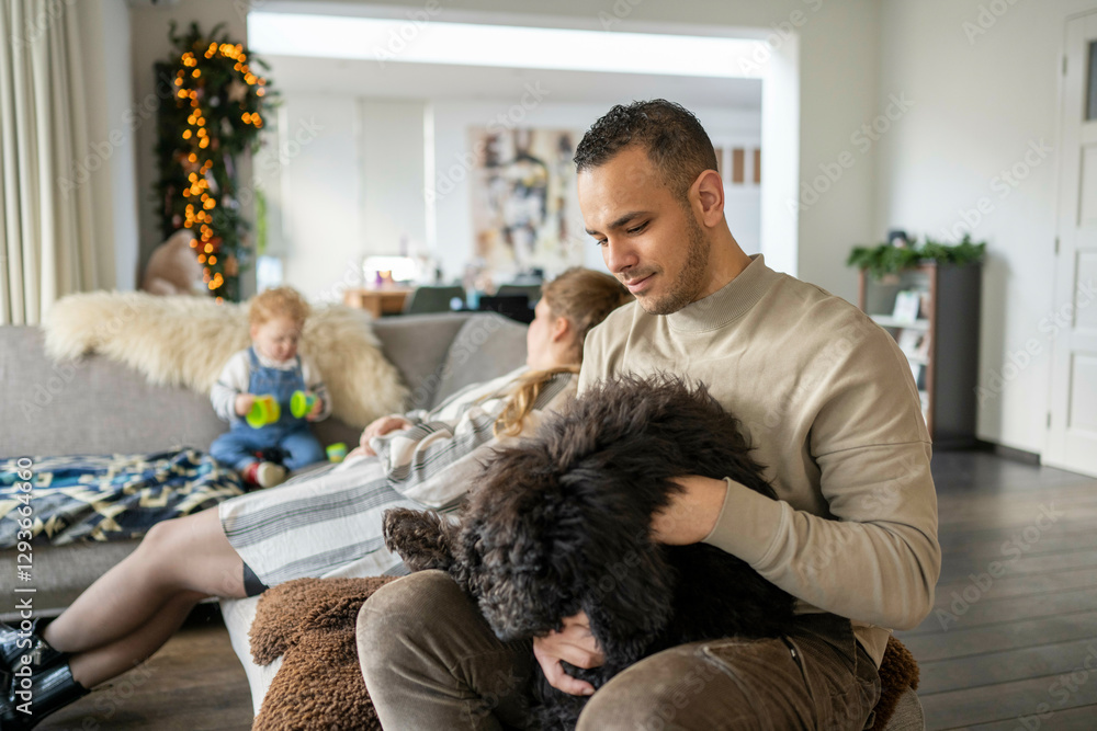 A family enjoys time at home with a father cuddling a dog and a child playing in the background, Netherlands A family enjoys time at home with a father cuddling a dog and a child playing in the background, Netherlands