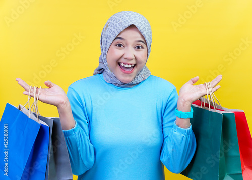 A cheerful asian woman in a hijab with shopping bags enjoys a fun shopping experience against a bright yellow background.