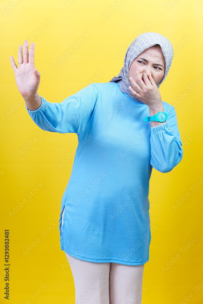 Naklejka premium An asian woman in a hijab and blue outfit holds her nose in disgust, reacting to a bad smell against yellow background.