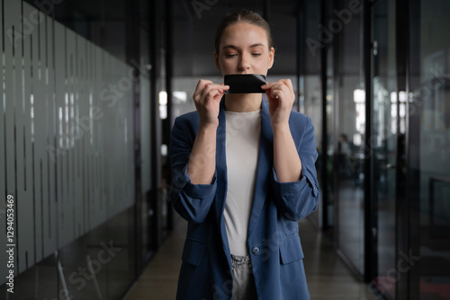 Foto Young woman holds a piece of black tape in front of her mouth, symbolizing censorship, silence, or restriction of speech in a modern office environment