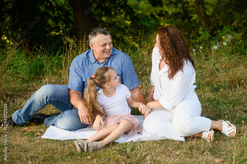 Family sitting happily together on cozy blanket spread on green grass. Mom, dad and daughter enjoying time outdoors spent together, talking, laughing. Happy family concept, vacation, family values. Family sitting happily together on cozy blanket spread on green grass. Mom, dad and daughter enjoying time outdoors spent together, talking, laughing. Happy family concept, vacation, family values.