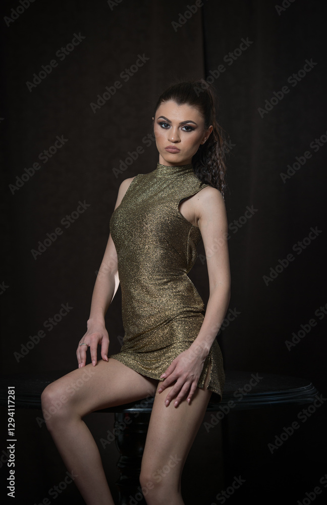 Portrait of a beautiful young brunette girl in a gold short dress on a dark background.