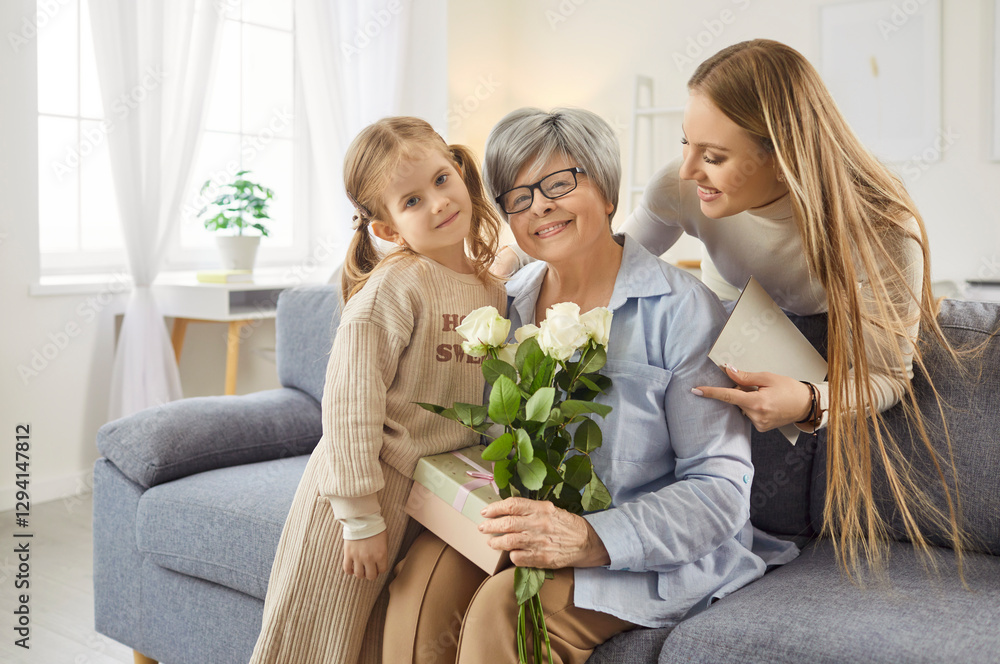 Smiling senior woman sitting on sofa, holding bouquet of white roses and gift box. Loving daughter and granddaughter standing nearby, expressing affection and celebrating special occasion at home. Smiling senior woman sitting on sofa, holding bouquet of white roses and gift box. Loving daughter and granddaughter standing nearby, expressing affection and celebrating special occasion at home.