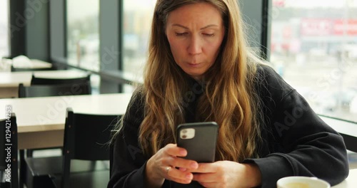 WARSAW, POLAND - MARCH 10 2023: Woman with long loose blonde hair sits in modern cafe and scrolls internet in smartphone. Cup of coffee stands in front of female focusing on screen