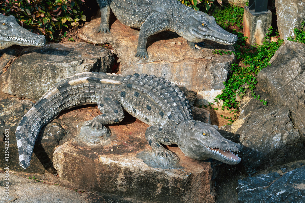 Fototapeta premium Beautiful realistic alligator stone statue decoration on stone near water. Large alligator statue laying on stone floor. Lifelike crocodile statue rests on rock by the riverbank as garden decoration.