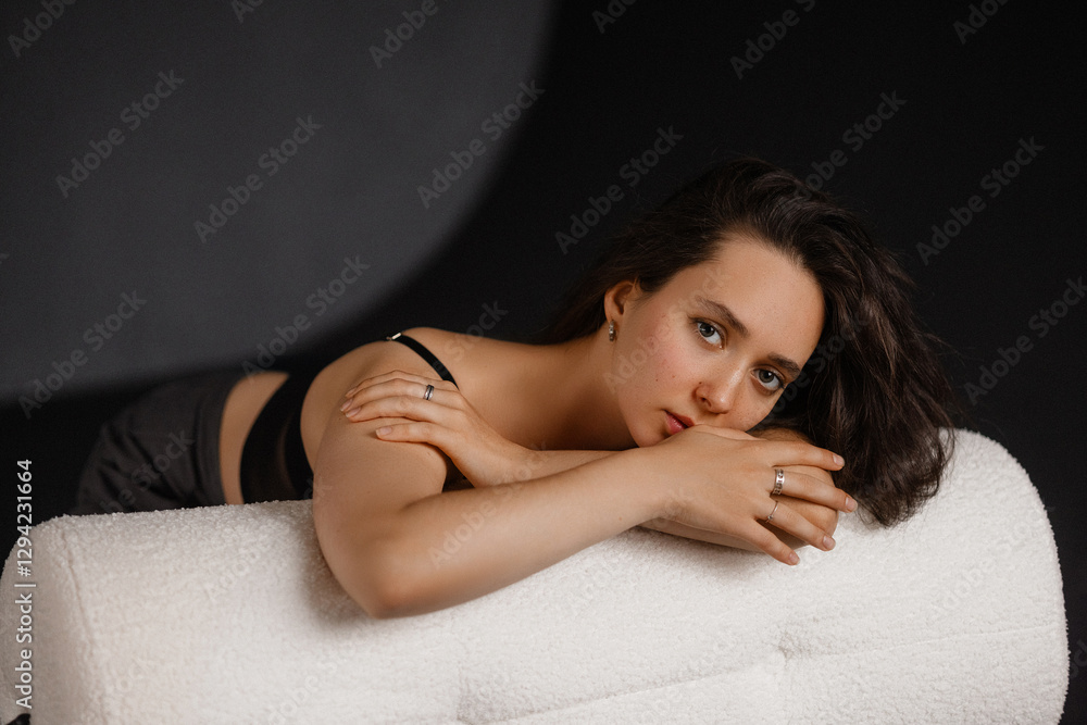 Naklejka premium A young Caucasian woman with dark hair reclines on a white couch against a dark background. Model caucasian in black top and gray trousers. Ideal for fashion, beauty, and relaxation concepts.