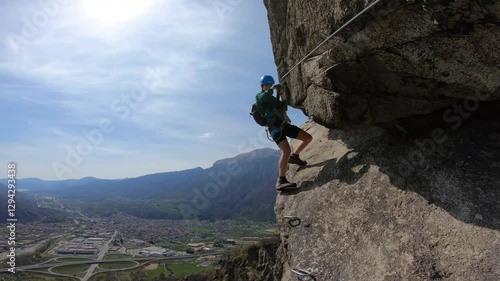 Via Ferrata Montorfano, Mergozzo: A lone climber braces against the jagged cliffside, carefully stepping along an exposed rock ledge with a breathtaking aerial view of the valley.