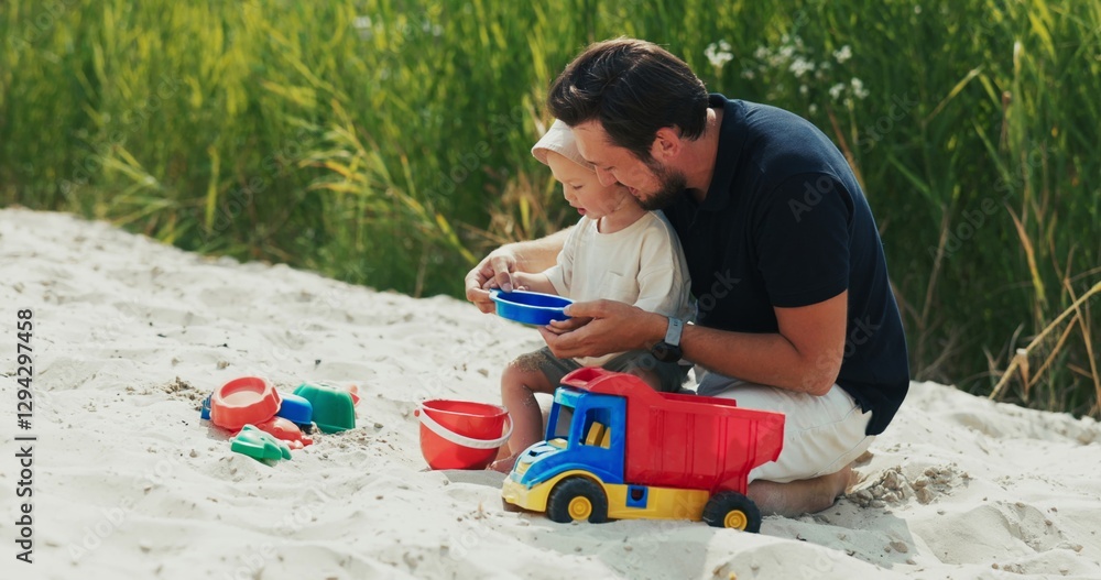 Naklejka premium On the beach, dad holds his son on his lap, teaching him to pour dry sand from a strainer into a bucket, surrounded by toys in the sandbox.