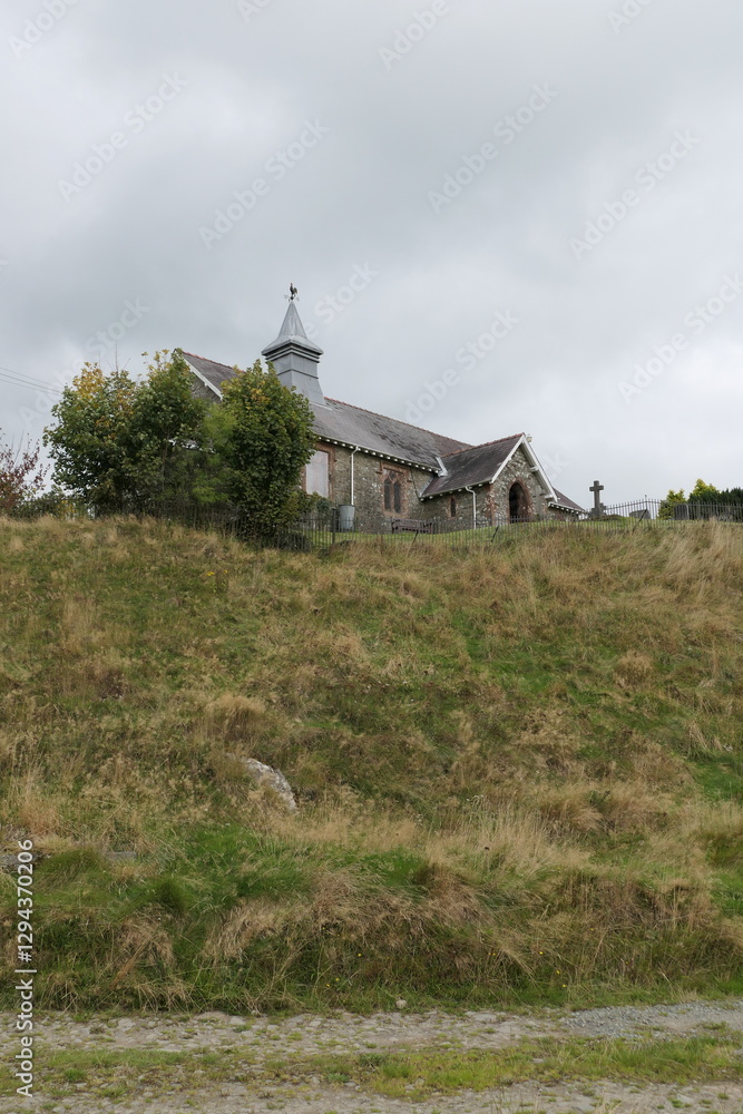 Obraz premium A small, charming stone church sits atop a gentle hill, surrounded by lush green fields in the Welsh countryside. Its weathered walls and modest bell tower stand against a backdrop of rolling hills.