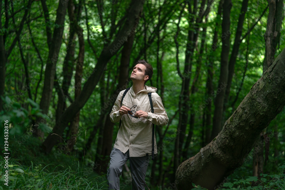 Adventurous man hiking in a forested mountain area, looking up while holding sunglasses. Sunlight filters through lush green trees. Adventurous man hiking in a forested mountain area, looking up while holding sunglasses. Sunlight filters through lush green trees.