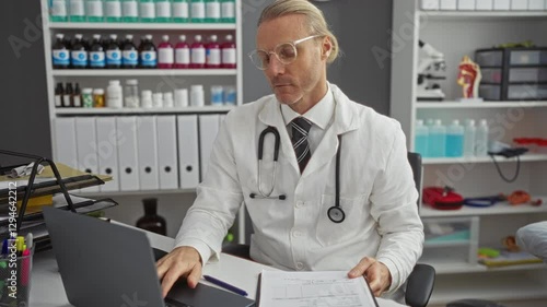 Wallpaper Mural Doctor in clinic reviews medical files on laptop in a professional office setting, with shelves of supplies in the background offering a glimpse of healthcare environment. Torontodigital.ca