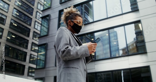 Wallpaper Mural Young stylish African-American businesswoman in a medical mask on her face, dressed in a business suit, is typing on a mobile phone, standing near a modern office building. Video in 4k, red komodo Torontodigital.ca