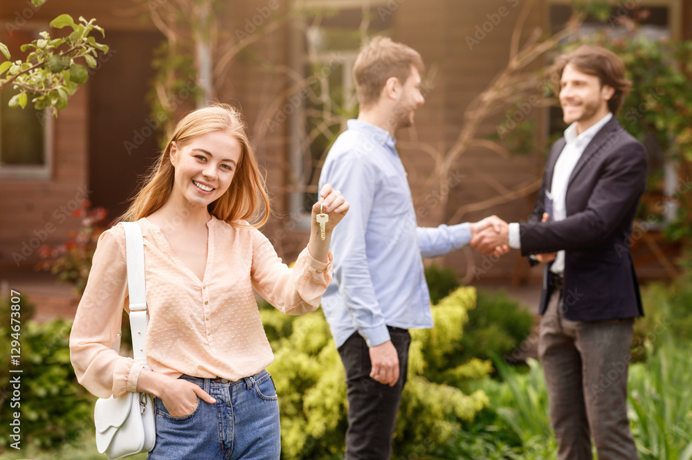 Happy young girl holding house key while her husband shaking hands with real estate agent outside Happy young girl holding house key while her husband shaking hands with real estate agent outside