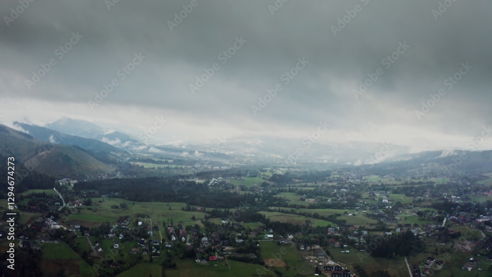 Fototapeta premium Vertical panning from a drone, a view of mountain valley with city in a lowland at foot of gently sloping mountains overgrown with a spruce forest, on a cloudy day at dusk
