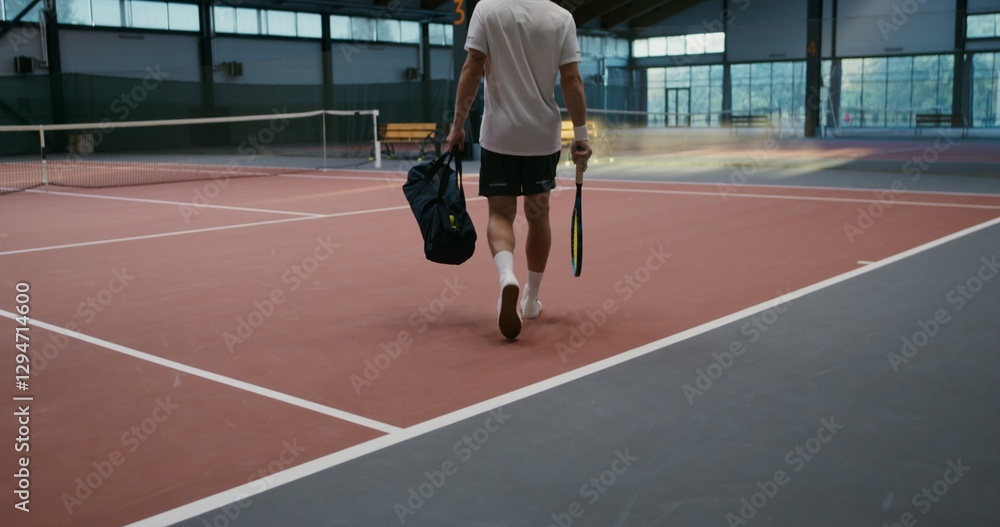 Fototapeta premium A young man in a sports uniform walks into an indoor tennis court with a racket and a bag of things, going to play tennis, video filming from the back