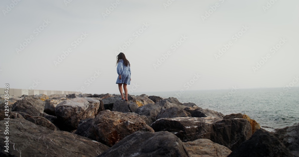 A young woman with flowing curly dark hair walks over large boulders, stepping from stone to stone, along the coastline on a clear summer day. 4k video, red komodo A young woman with flowing curly dark hair walks over large boulders, stepping from stone to stone, along the coastline on a clear summer day. 4k video, red komodo