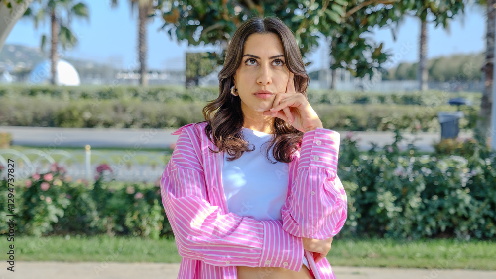 A young brunette businesswoman in a tree-filled park on a spring day, looking at the camera with a thoughtful expression A young brunette businesswoman in a tree-filled park on a spring day, looking at the camera with a thoughtful expression