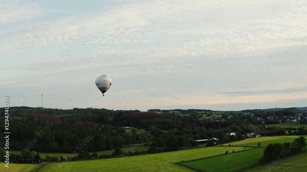 Obraz premium Drone shot of a hot air balloon in the sky flying over a green valley near a village in Germany on a sunny day.