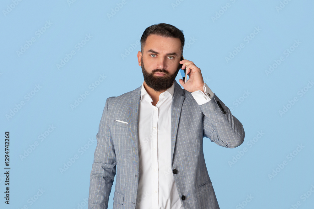 Serious millennial businessman in office wear having conversation on smartphone on blue studio background. Young bearded CEO making phone call, speaking on mobile device