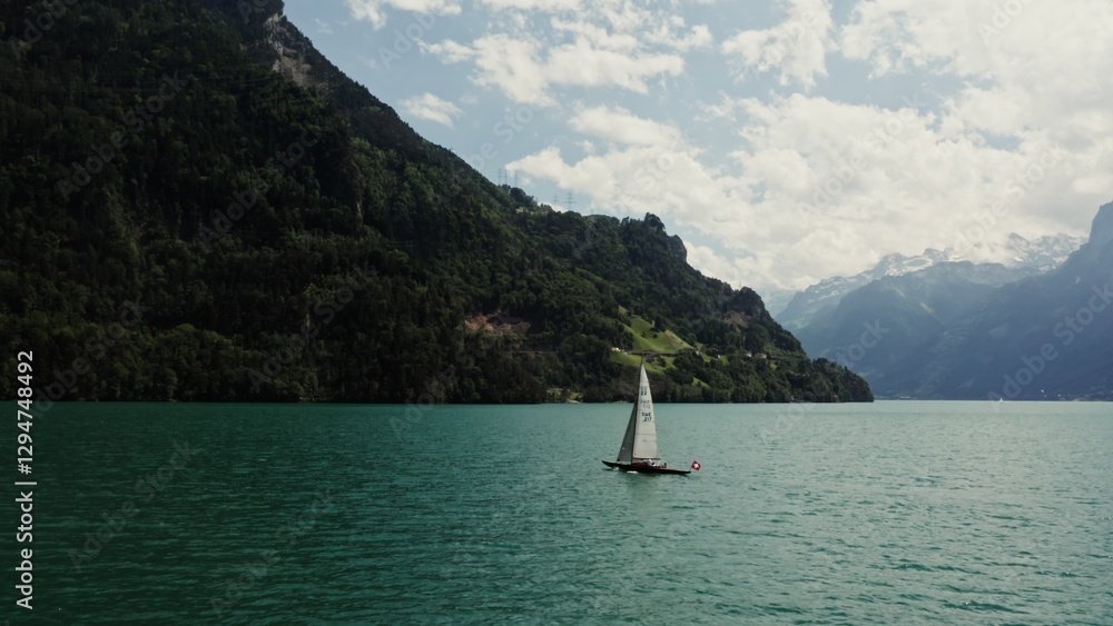 Naklejka premium A sailing boat with a Swiss flag floats on a picturesque lake at the foot of the Alps, the tops of which are covered with white clouds. Panoramic drone footage, Switzerland