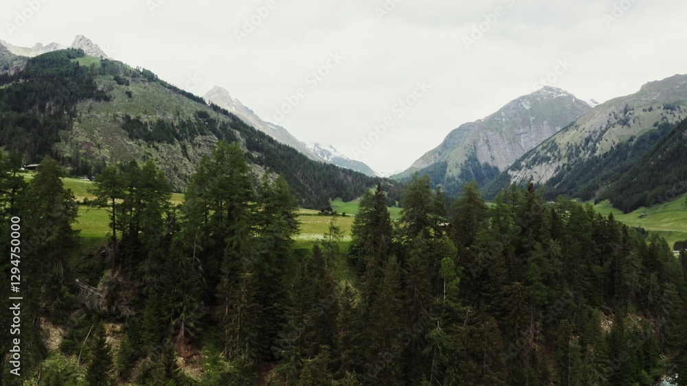 The drone rises from behind the trees, revealing beautiful panoramic view of high mountain valley with a village in the lowland. The peaks of the rocky Alpine mountains are covered with snow. Austria The drone rises from behind the trees, revealing beautiful panoramic view of high mountain valley with a village in the lowland. The peaks of the rocky Alpine mountains are covered with snow. Austria