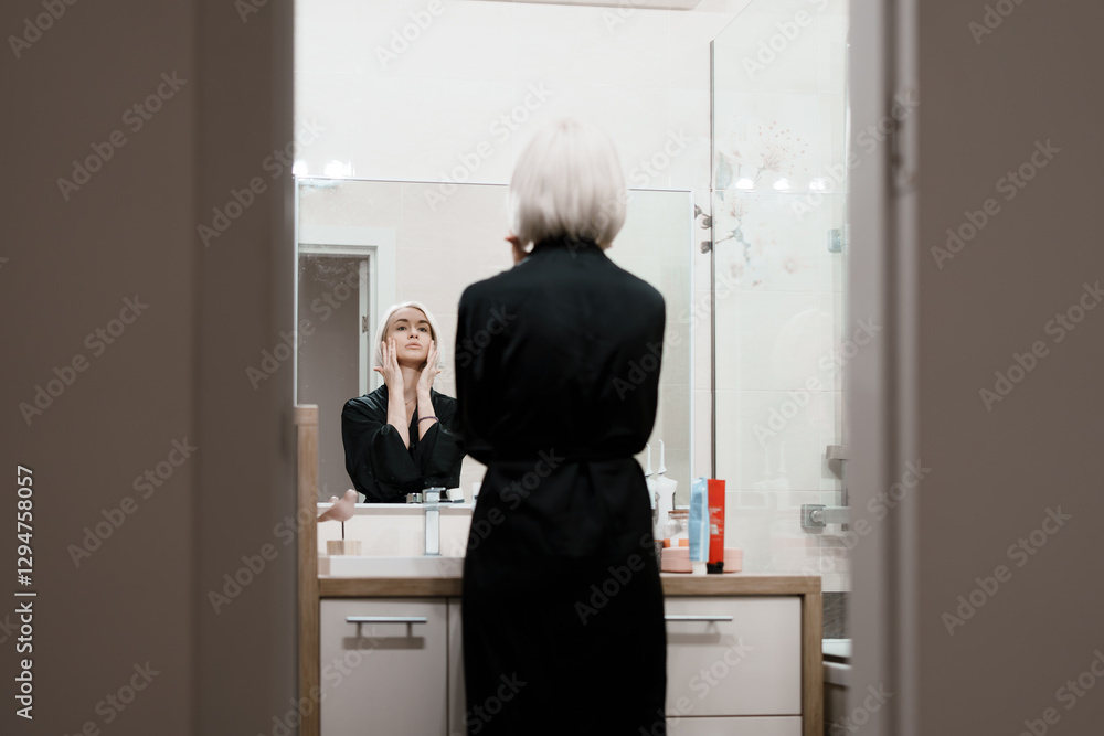 A woman in a black robe stands in front of a bathroom mirror, applying skincare products as part of her daily beauty routine in a well-lit modern bathroom. A woman in a black robe stands in front of a bathroom mirror, applying skincare products as part of her daily beauty routine in a well-lit modern bathroom.