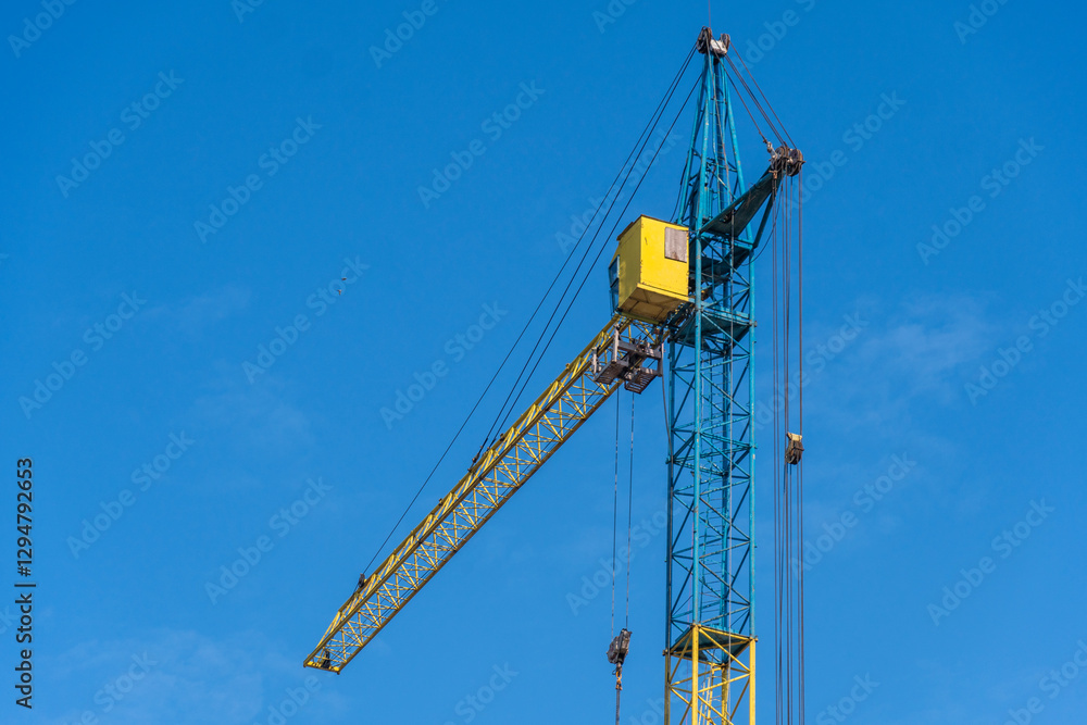 A large construction crane stands tall against a vibrant blue sky, its yellow and blue structure clearly visible under the bright sunlight in the daytime
