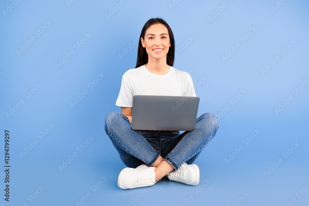 Naklejka premium Technology. Portrait of a smiling young woman sitting with legs crossed, using laptop computer isolated over blue studio background, looking at camera. Satisfied lady studying or working with notebook